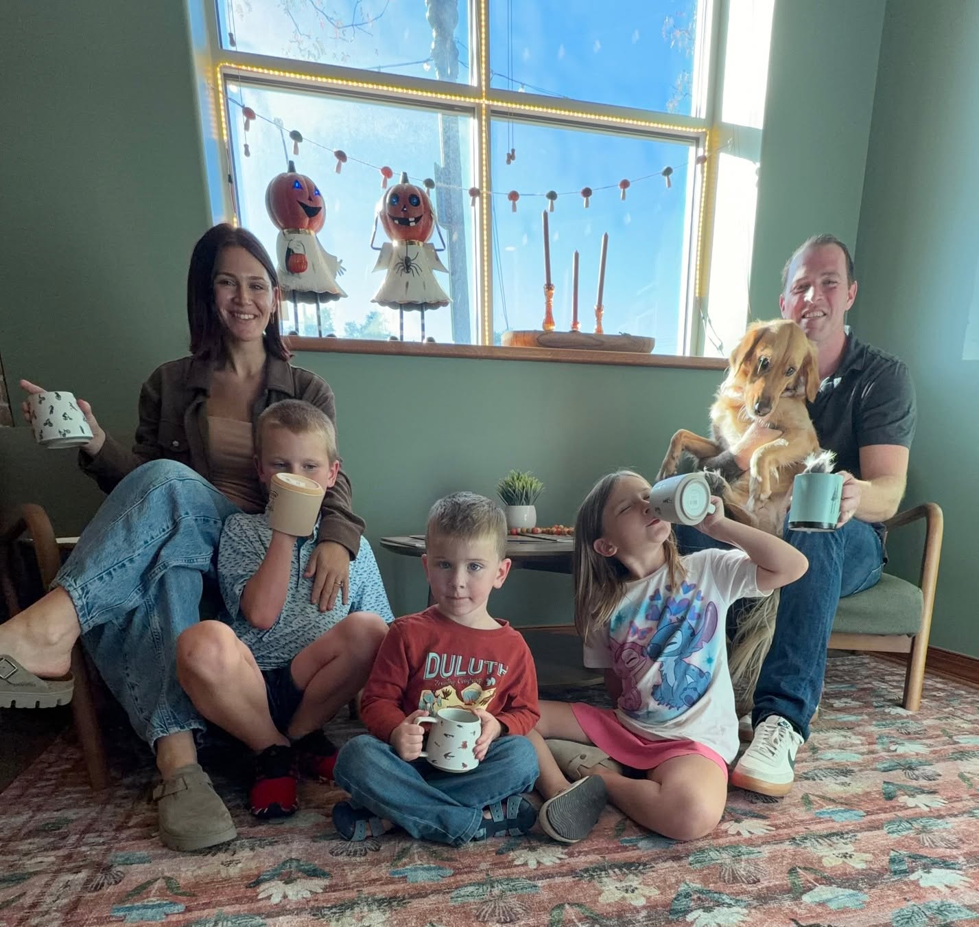 The Hummingbird Collective owners and their three children and golden dog sitting on the floor of the shop, each holding a coffee mug